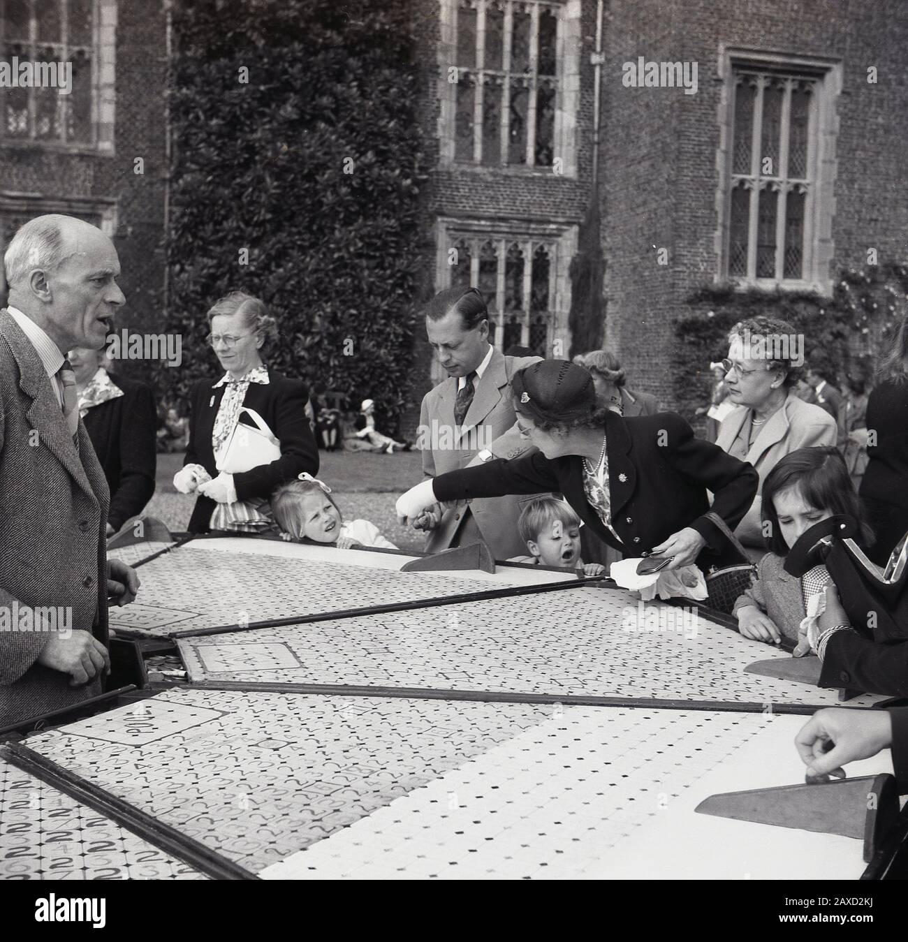 1950s, historical, in the grounds of a country house at a english summer garden party, well-dressed adults in the clothes of the day, with their children at a stall, playing a game of shove half penny or roll a penny, England, UK. A traditional game, often played in pubs and social clubs, it involves players pushing or rolling coins up a board. Stock Photo