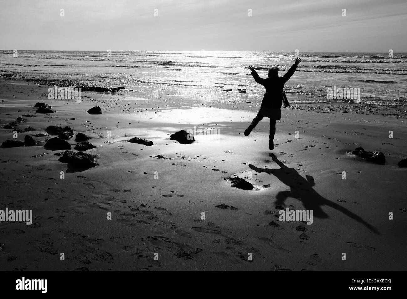 Woman jumps for joy on the beach in silhouette in strong late afternoon winter sun with strong shadow on sand Stock Photo
