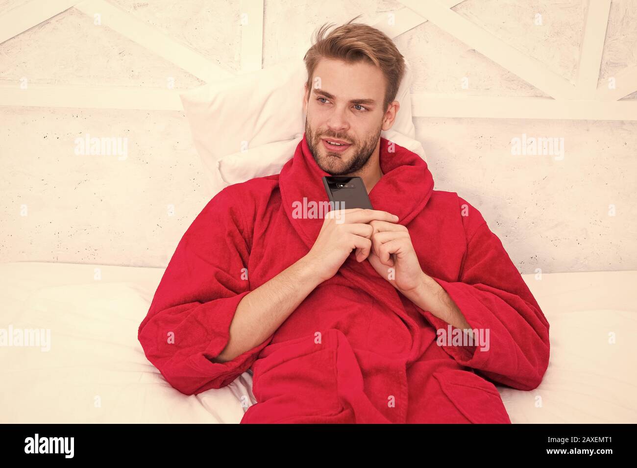 Creating his own blog. Handsome man making new blog post from smartphone. Blogger keeping private personal blog in bed. Caucasian guy in bathrobe posting blog on online social network from bedroom. Stock Photo