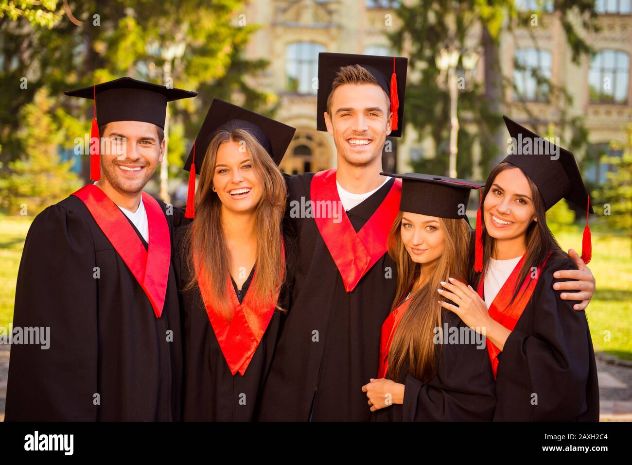 successful joyful five graduates in robes and hats smiling and hugging Stock Photo