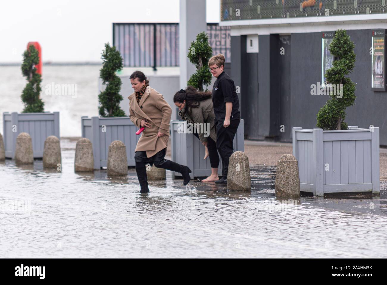 A female wades through flood water whilst another removes her shoes after being cut off by flooding during the high tide storm surge of Storm Ciara Stock Photo
