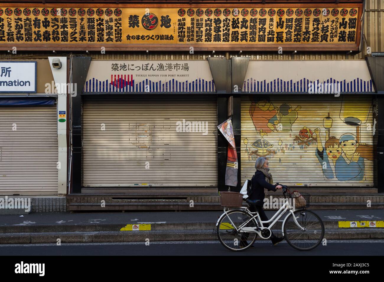 An older Japanese woman pushes a bicycle passed the shuttered store fronts of shops in what remains of Tsukiji outer market. Tsukiji, Tokyo, Japan. Stock Photo