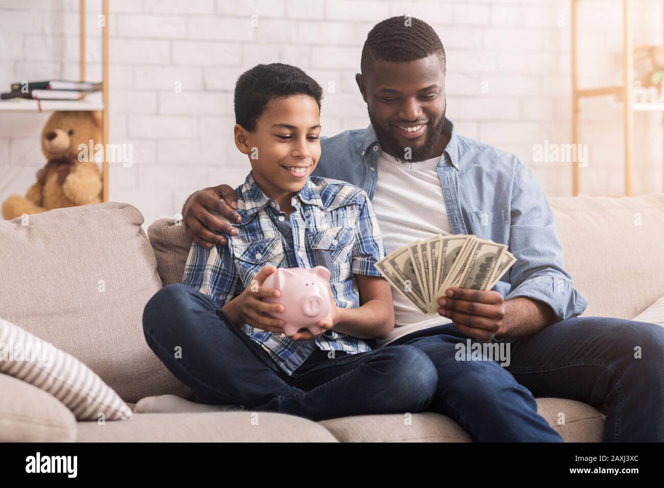 Personal Savings. Little Afro Girl Putting Money To Piggybank, Father Holding It Stock Photo