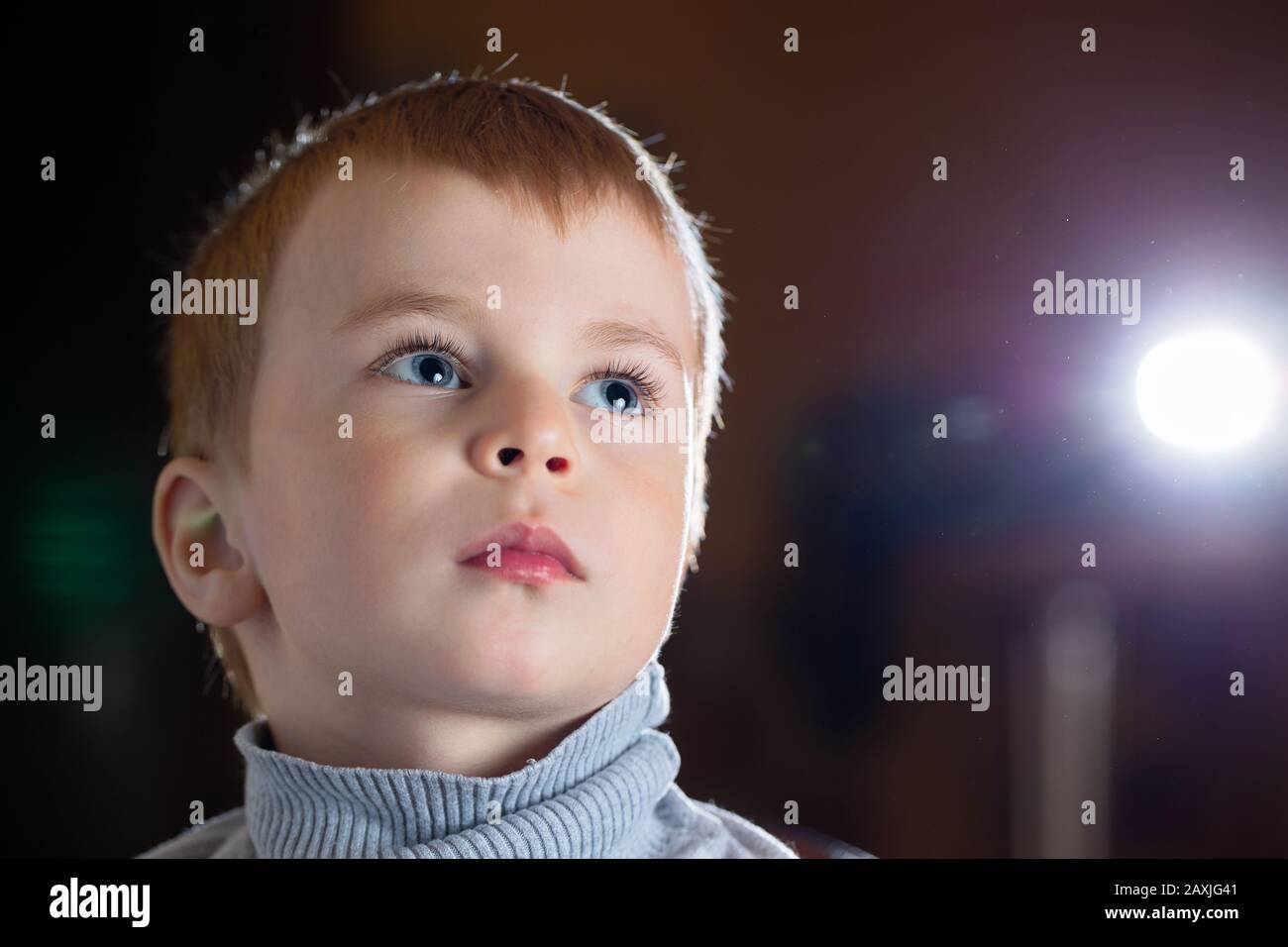 Facial portrait of dreamy pensive blond boy with gaze his eyes fixed upwards over dark background, with specks of dust flying in backlight with flares Stock Photo