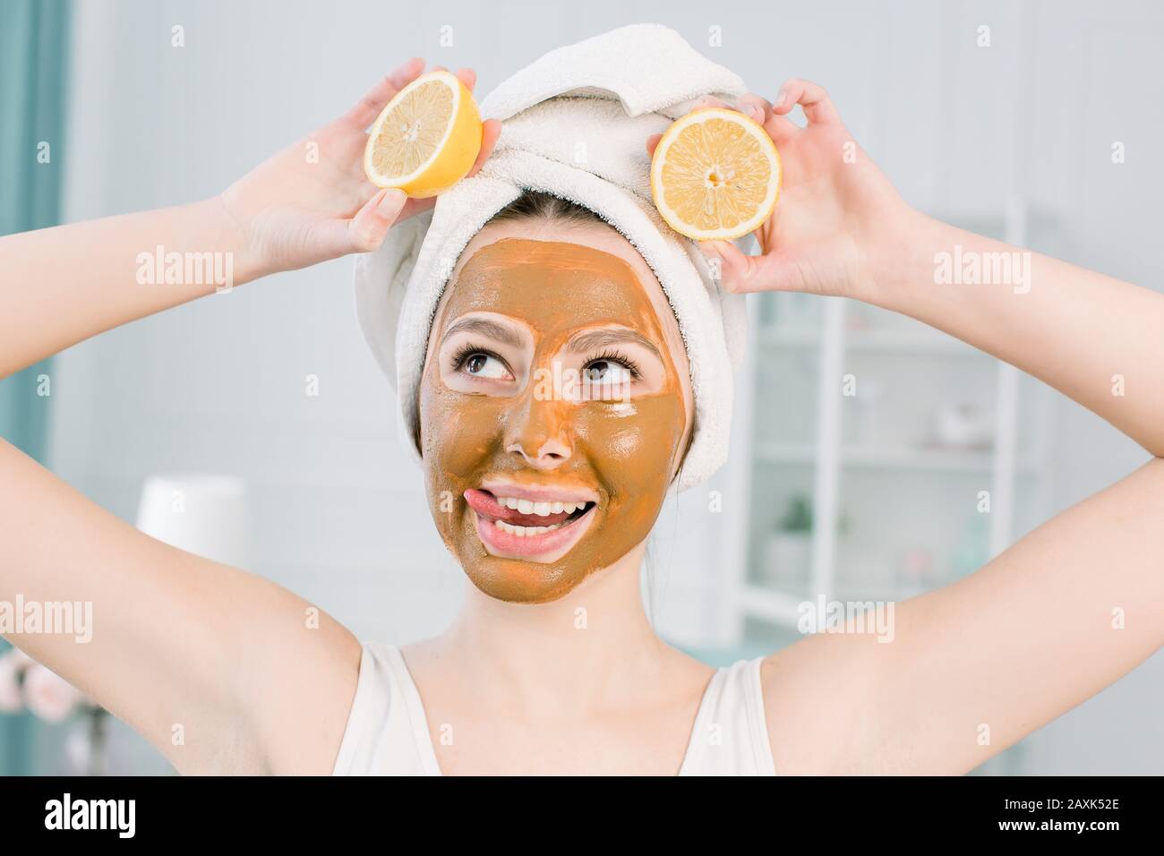 beautiful Caucasian woman in white towel with brown clay mask on her face and lemon slices, showing tongue and kiss face, beauty treatments concept Stock Photo
