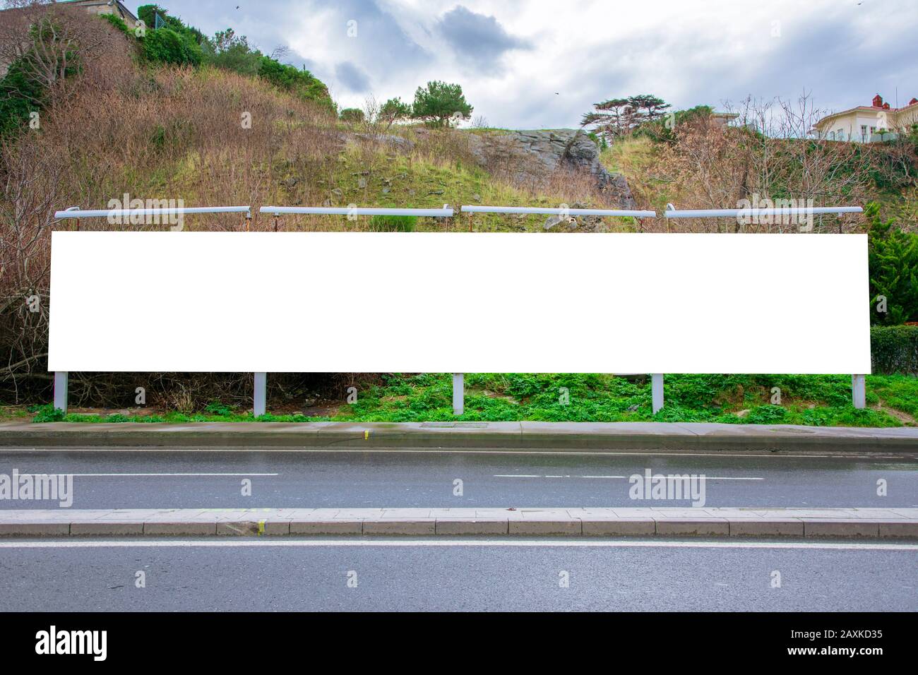 Blank billboard at the roadside photographed from the opposite sidewalk, with a deserted hill in the background Stock Photo