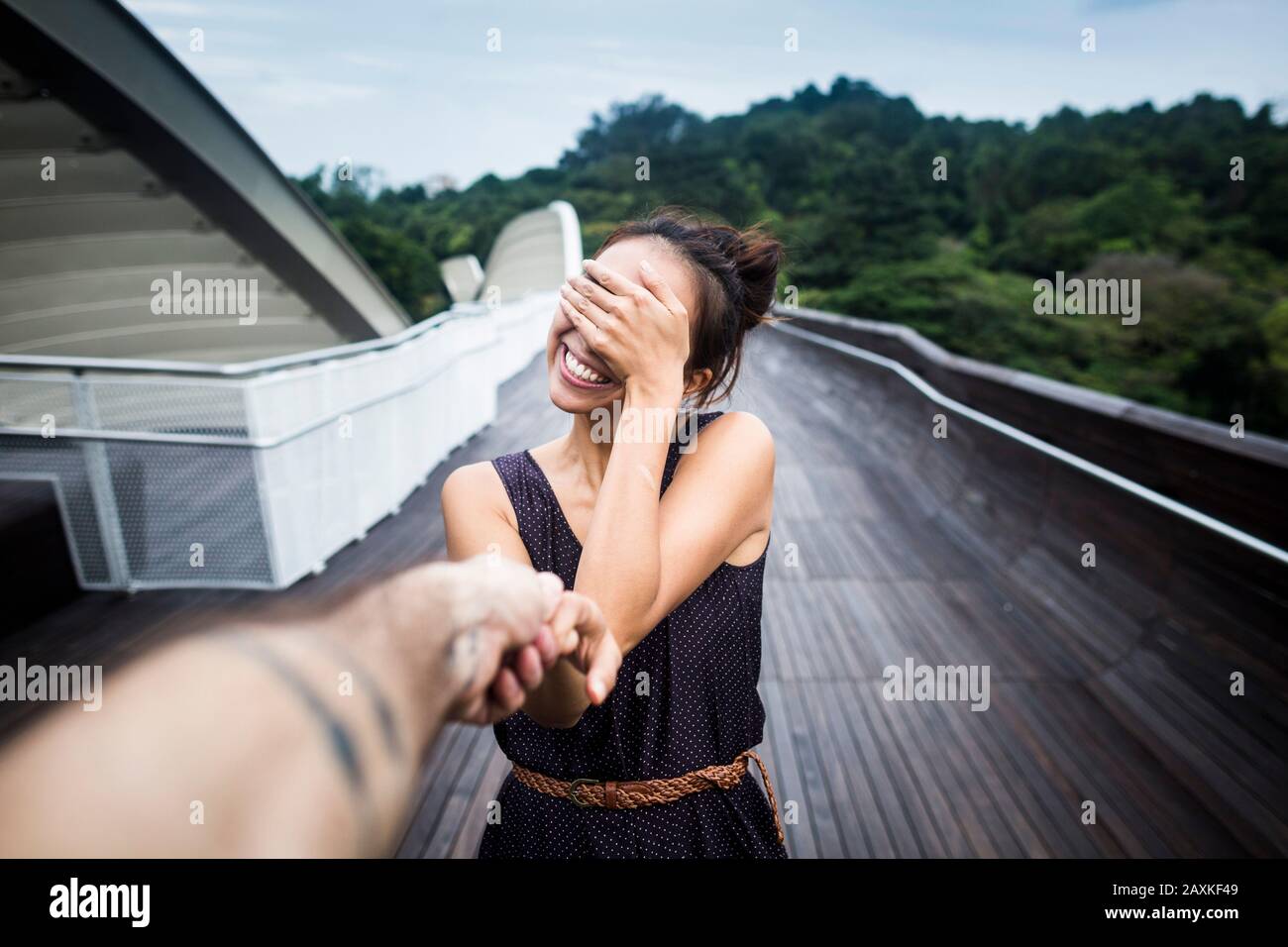 Smiling young woman standing on a bridge, covering her face, holding man's hand. Stock Photo
