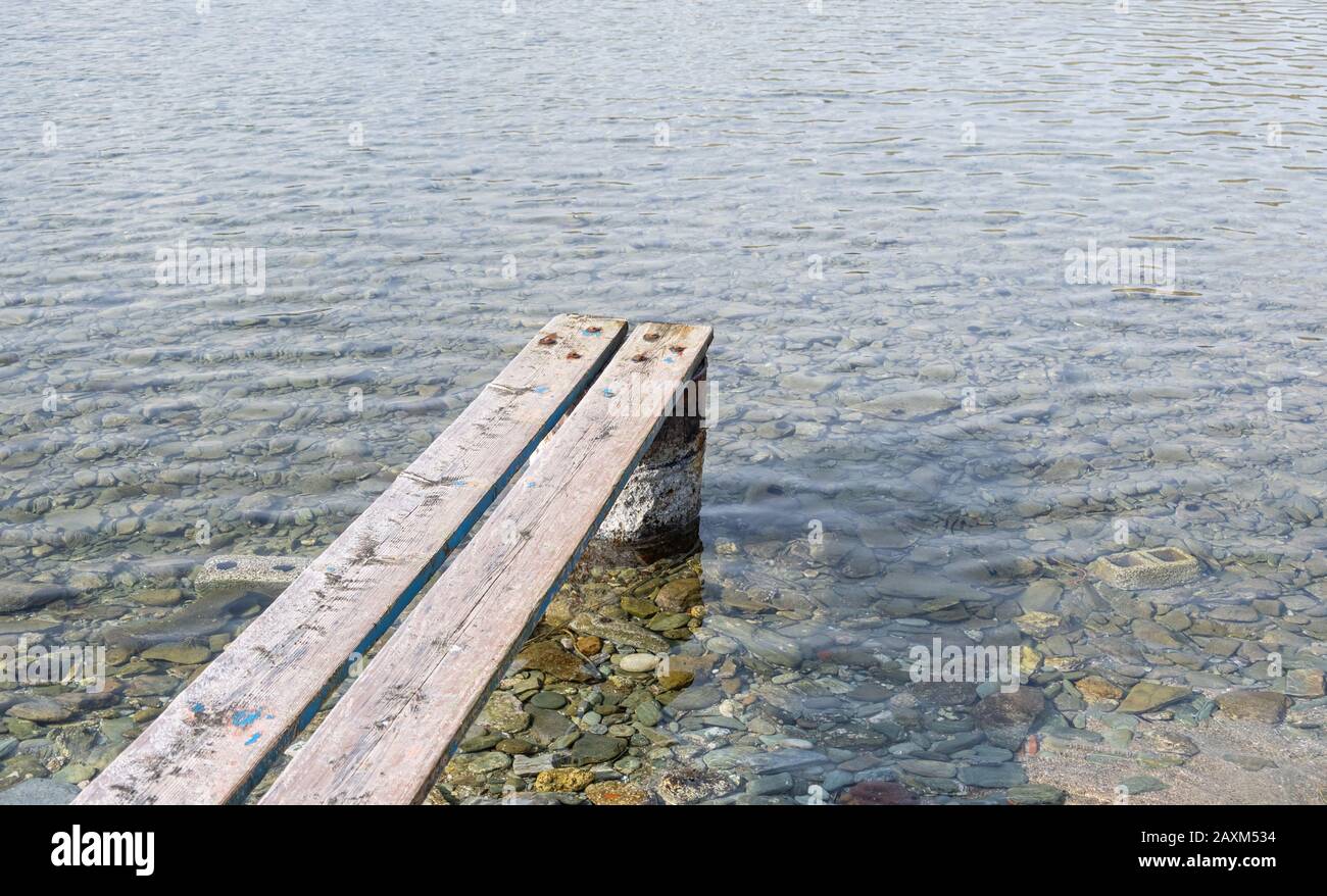 Weathered wooden small pier. Wood planks set properly on rusty barrel to make a mini platform over a transparent calm blue sea background. Stock Photo
