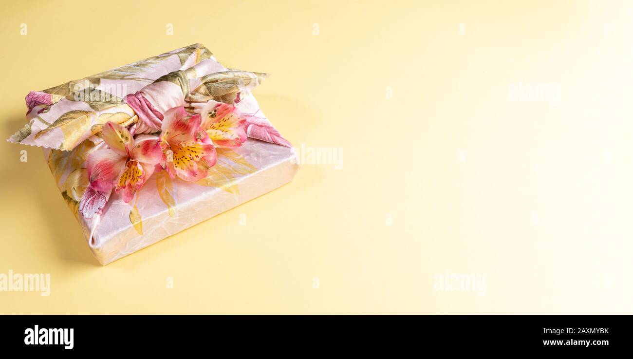 Gift box trendy wrapped in floral cloth in japanese Furoshiki technique with flowers Alstroemeria on yellow background. Valentine's day or Women's day Stock Photo