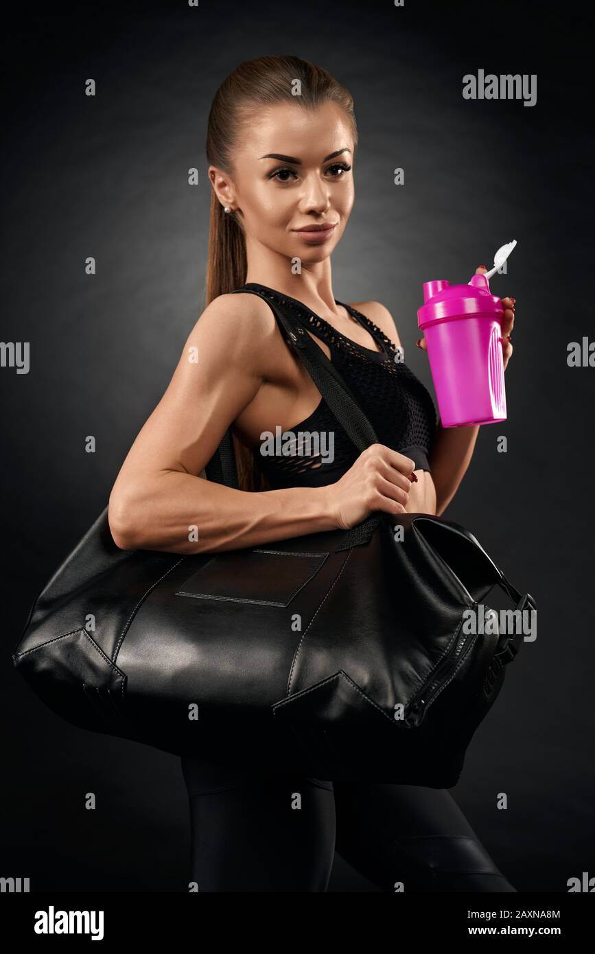 Young attractive fit woman posing with sport bag and holding pink shaker with water, isolated on black. Portrait of pretty female with ponytail in black sportswear going home after hard training. Stock Photo