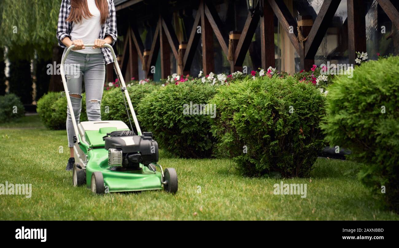 Horizontal crop of woman in casual outfit using lawn mower on backyard. Selective focus of young gardener working in summer, cutting grass near bushes in backyard. Concept of gardening, work, nature. Stock Photo