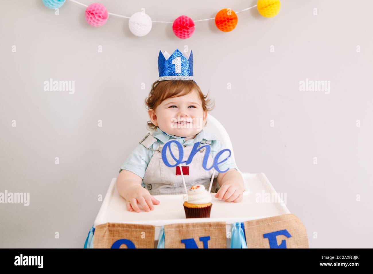 Cute adorable Caucasian baby boy in blue crown celebrating his first birthday at home. Child kid toddler sitting in high chair eating tasty cupcake de Stock Photo