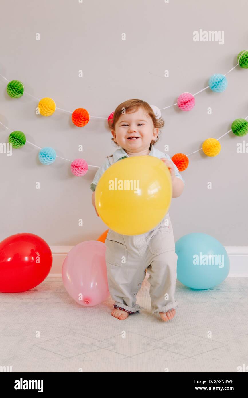 Funny Caucasian baby boy celebrating his first birthday. Excited child kid toddler playing with colorful balloons. Celebration of event or party indoo Stock Photo