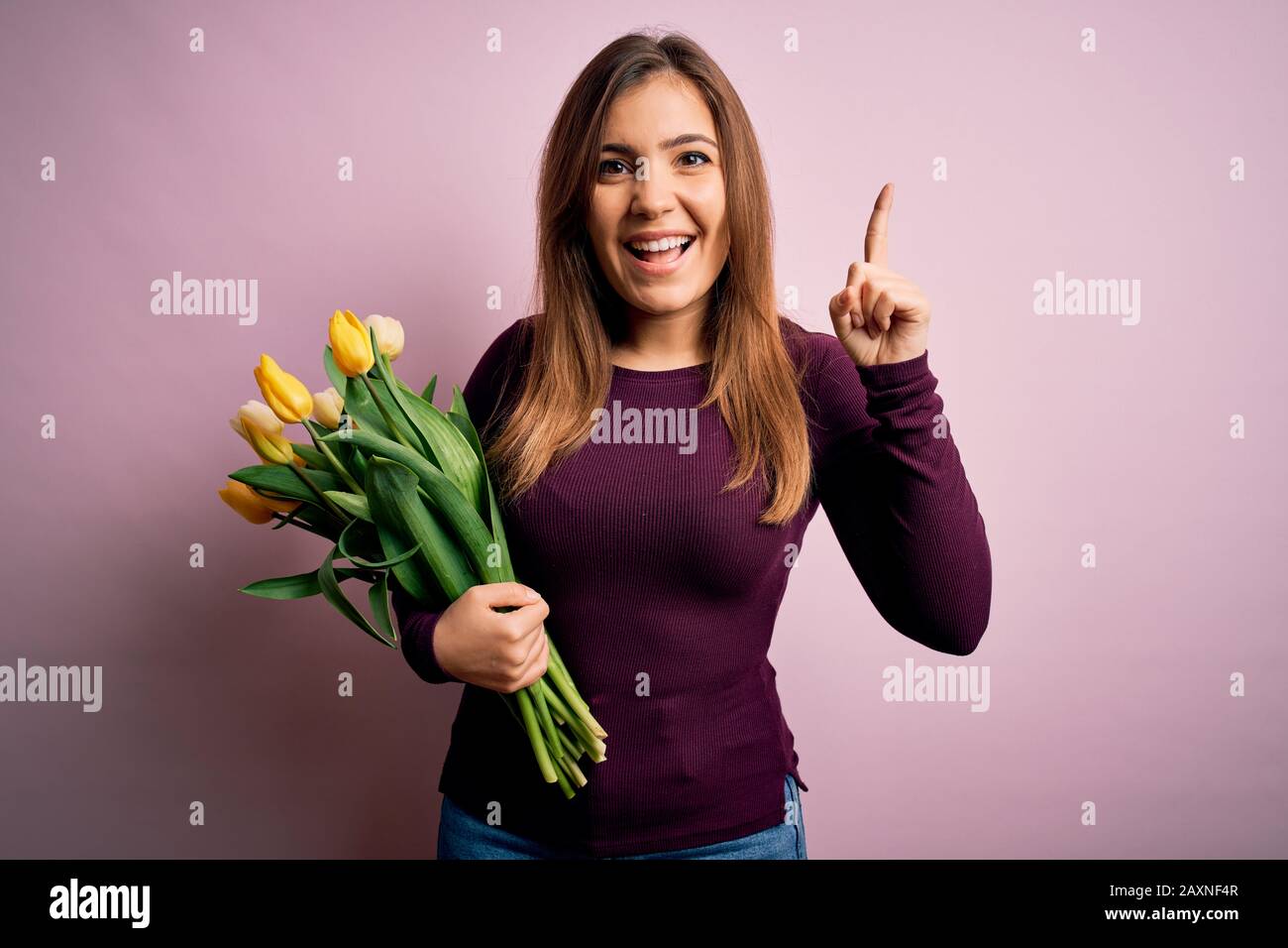 Young blonde woman holding romantic bouquet of yellow tulips flowers over pink background smiling amazed and surprised and pointing up with fingers an Stock Photo