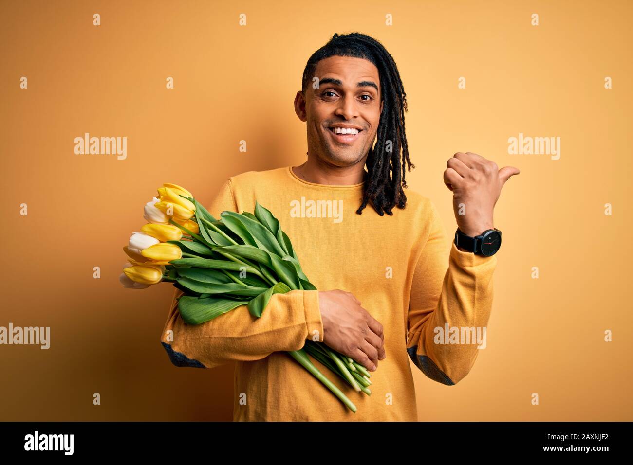 Young african american afro romantic man with dreadlocks holding bouquet of yellow tulips smiling with happy face looking and pointing to the side wit Stock Photo