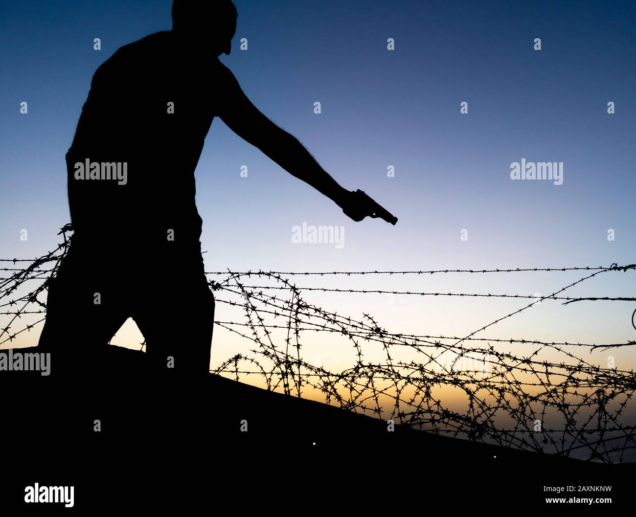 Silhouette of man pointing gun near barbed wire fence. Stock Photo