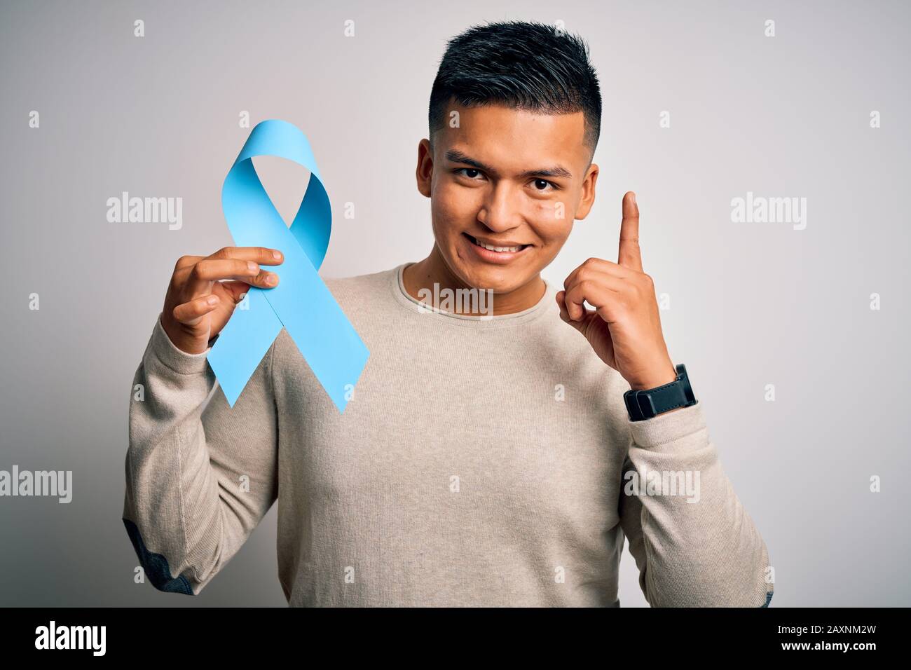 Young handsome latin man holding blue cancer ribbon over isolated white ...