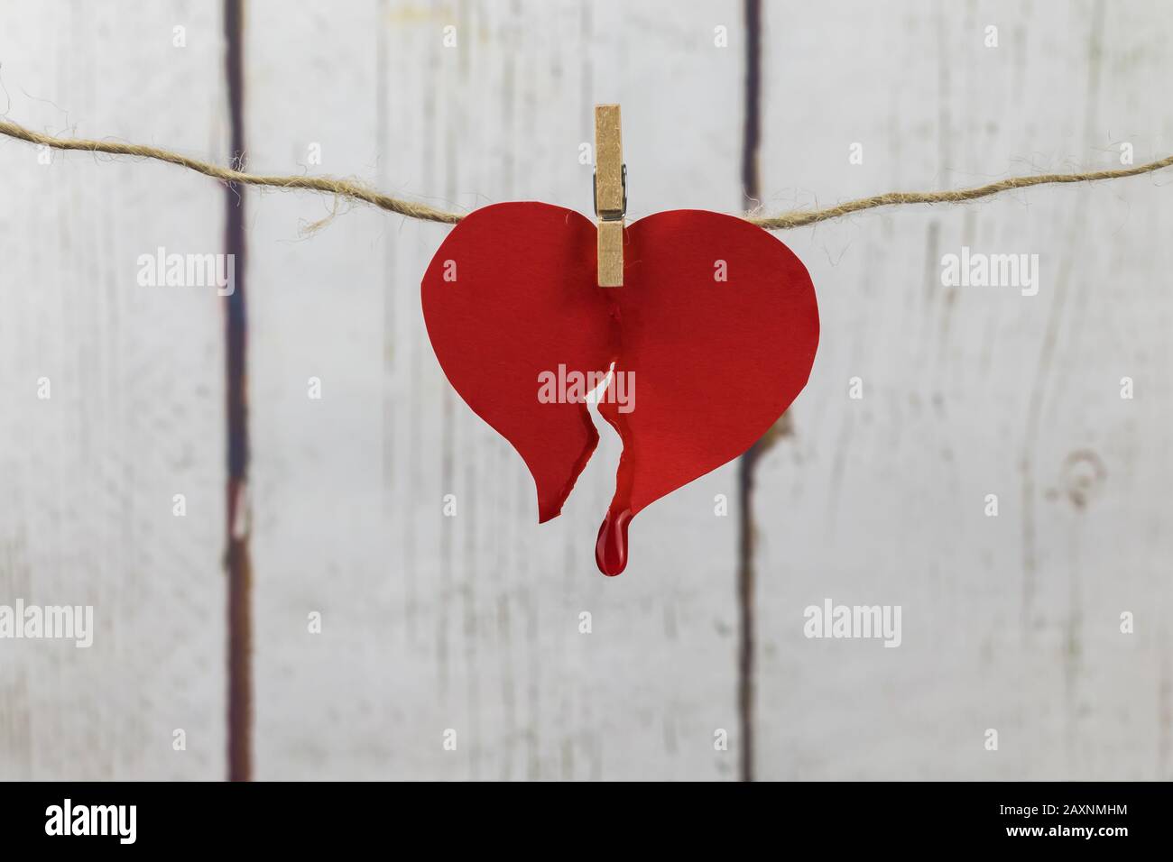 Broken heart of red cardboard with blood drop, hung with tweezers on a white wooden background Stock Photo