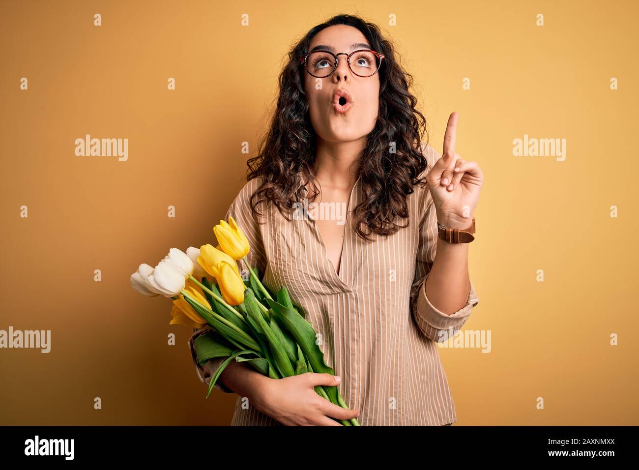 Young beautiful romantic woman with curly hair holding bouquet of yellow tulips amazed and surprised looking up and pointing with fingers and raised a Stock Photo