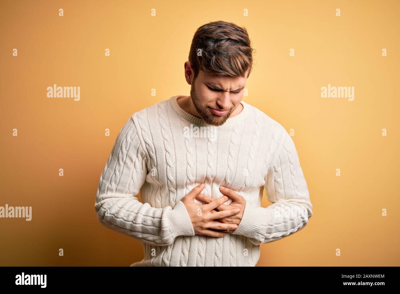 Young blond man with beard and blue eyes wearing white sweater over yellow background with hand on stomach because indigestion, painful illness feelin Stock Photo