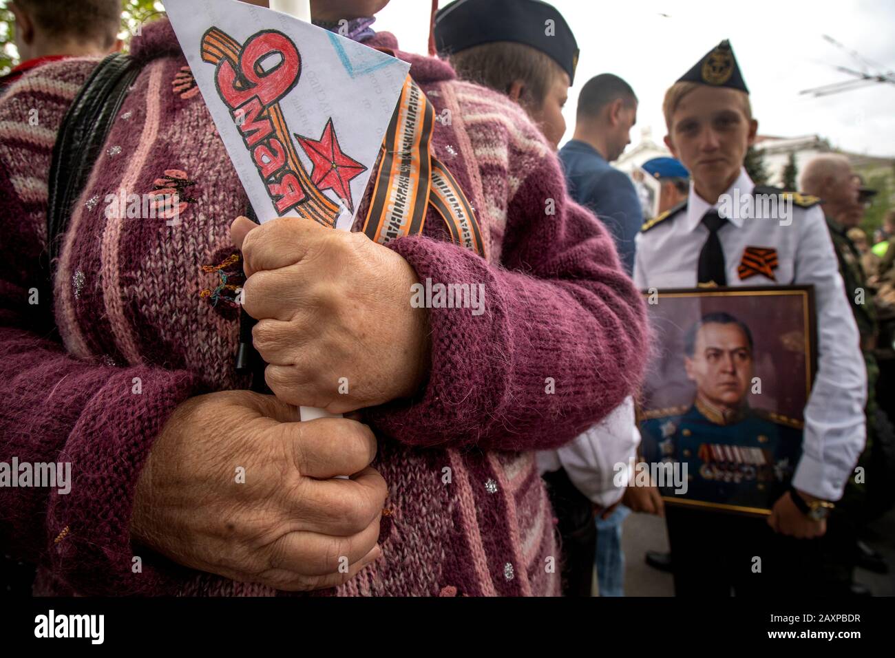 Sevastopol, Crimea, 9th of May, 2019 Old woman holds a portrait with photography of her relatives, of those killed in World War II, and a postcard with inscription in Russian 'The 9 May' during the action he Immortal Regiment in Sevastopol city. A boy in the background holds a portrait of Marshal of Soviet Union Georgy Zhukov Stock Photo