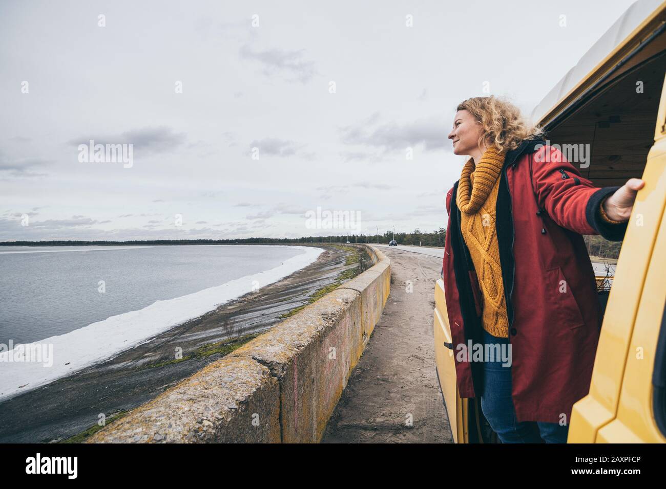 Young blond woman looking out of camper van overlooking the frozen winter sea Stock Photo