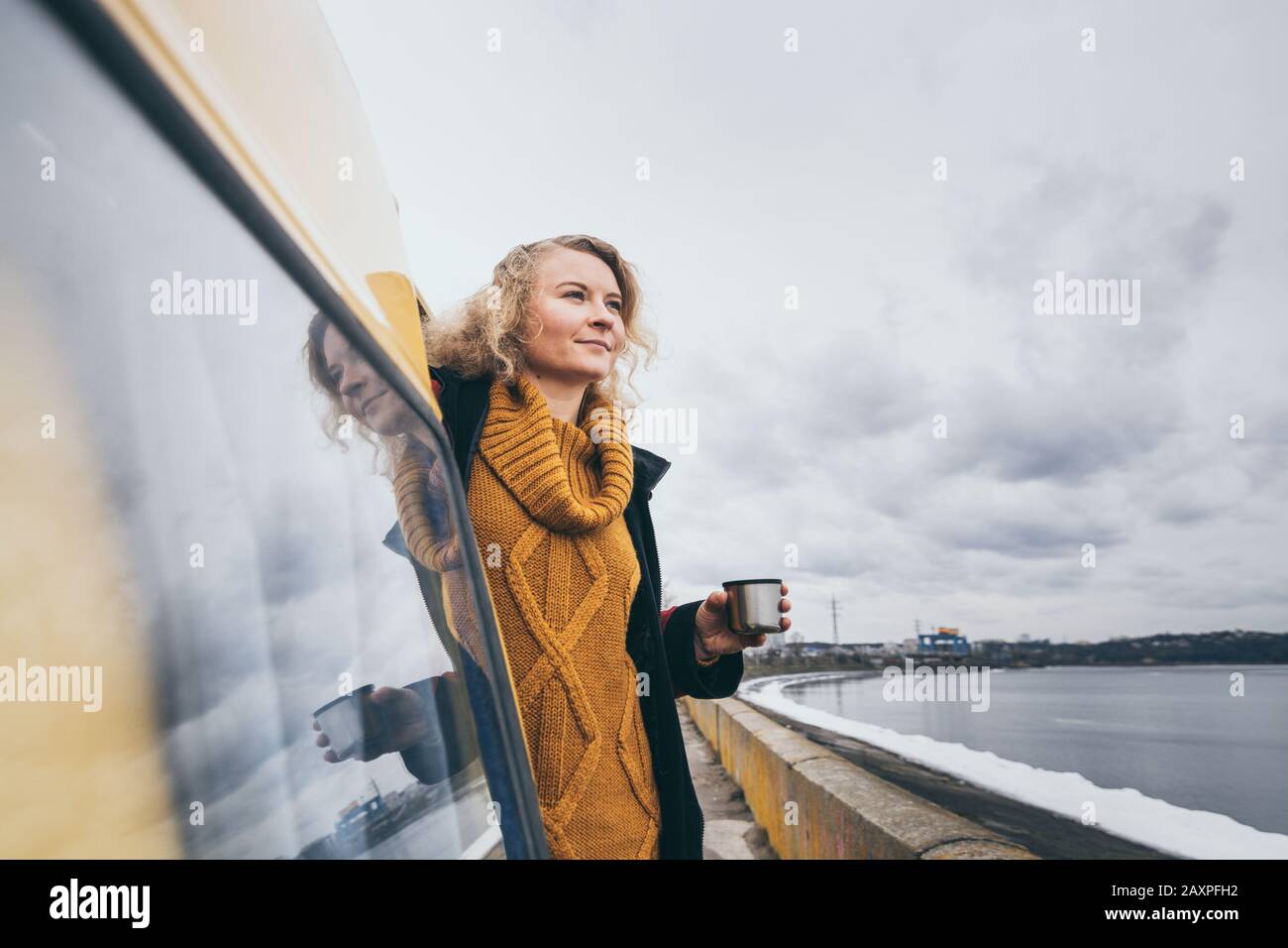 Young blond woman looking out of camper van overlooking the frozen winter sea. Reflection in the car window. Stock Photo