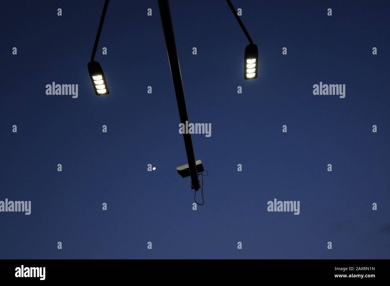 Street lights and surveillance cameras arm reaches across a deep blue sky observing the the moon