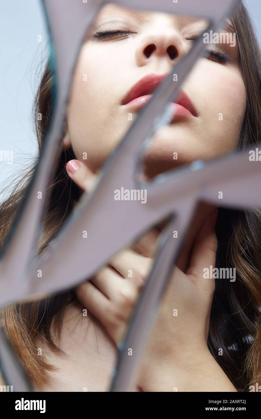 Young woman looks in a broken mirror. Portrait of beautiful female in the mirror shards. Hand on the throat. Stock Photo