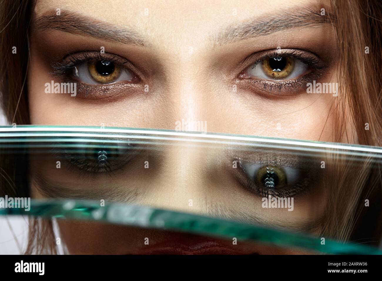 Portrait of a beautiful girl with a mirror splinter near eyes. Female with mirror shard in hand posing on gray background. Stock Photo