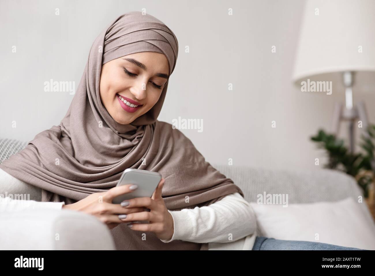 Cheerful arabic girl in hijab spending time with smartphone at home Stock Photo