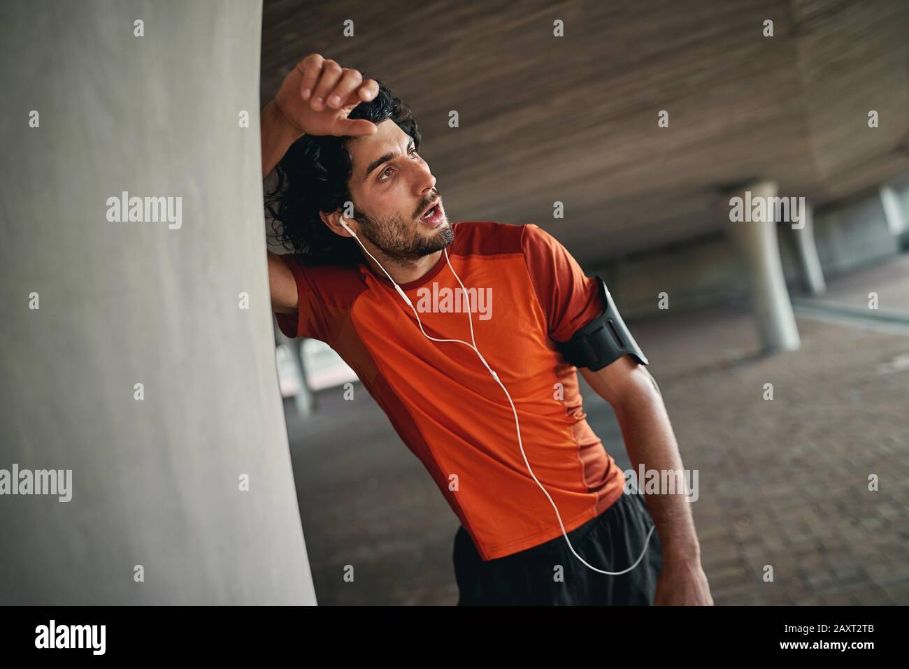 Exhausted sporty athletic young man with earphones and armband leaning on column under the bridge taking rest after running - exhausted athlete Stock Photo
