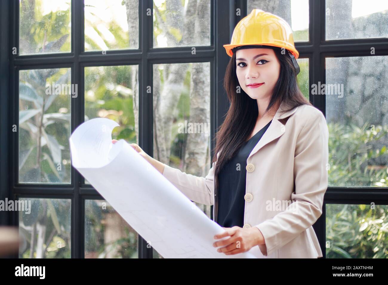 portrait of beautiful Asian woman architect builder with yellow helmet hard hat studying blueprint plan of the rooms, serious civil engineer working w Stock Photo