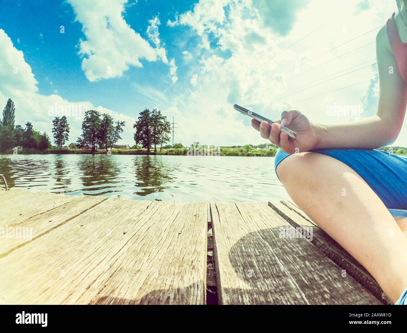 A girl with a phone in her hand sits on the wood surface near the surface of the water. Shot from below. Wide angle action camera go pro. Stock Photo