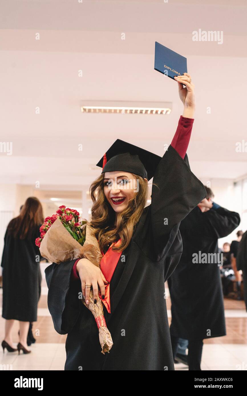 Young happy female graduate student wearing mortarboard holding diploma and bouquet of flowers while standing in university at graduation ceremony Stock Photo