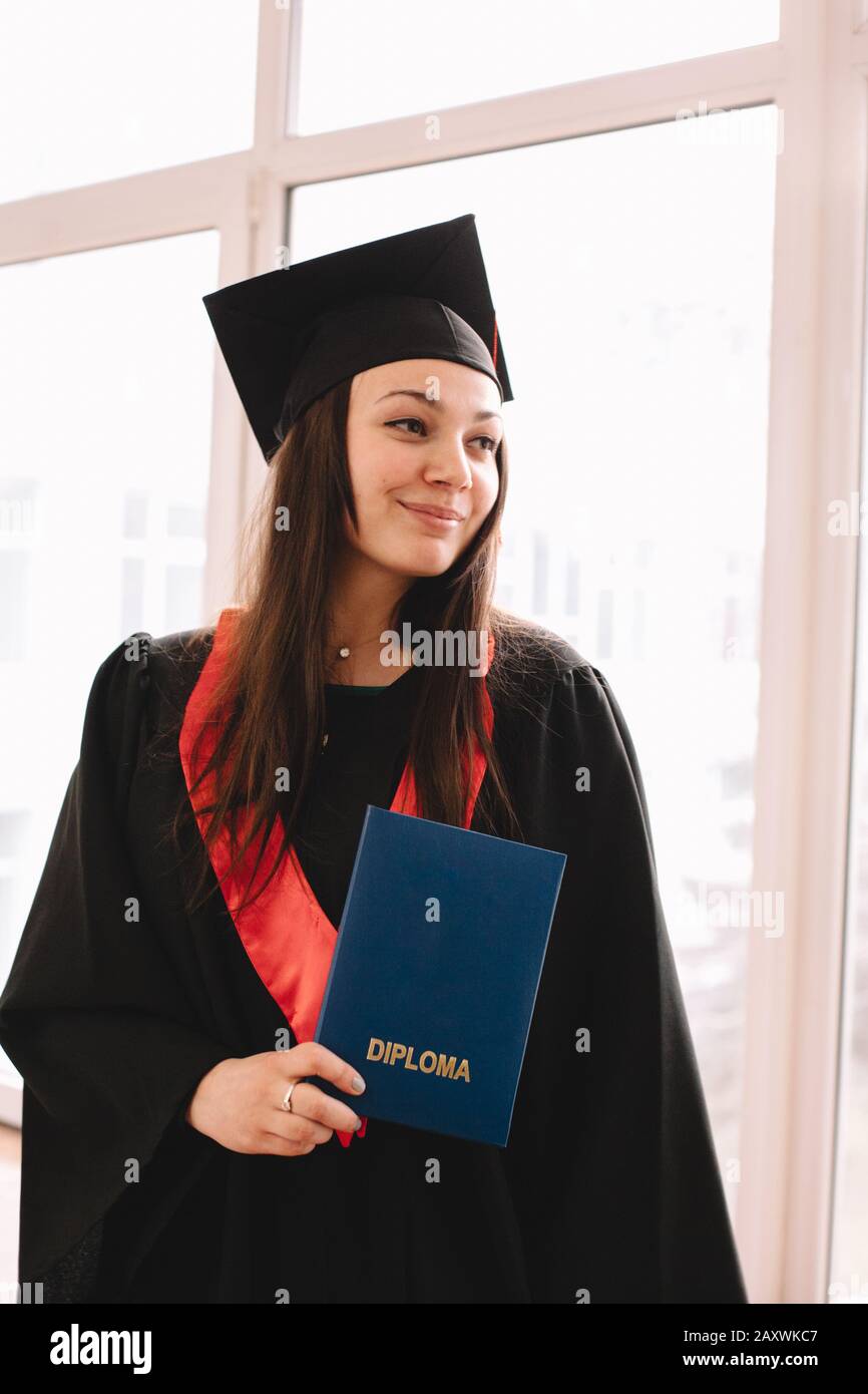 Portrait of happy young woman in graduation gown holding diploma while standing in university at graduation ceremony Stock Photo
