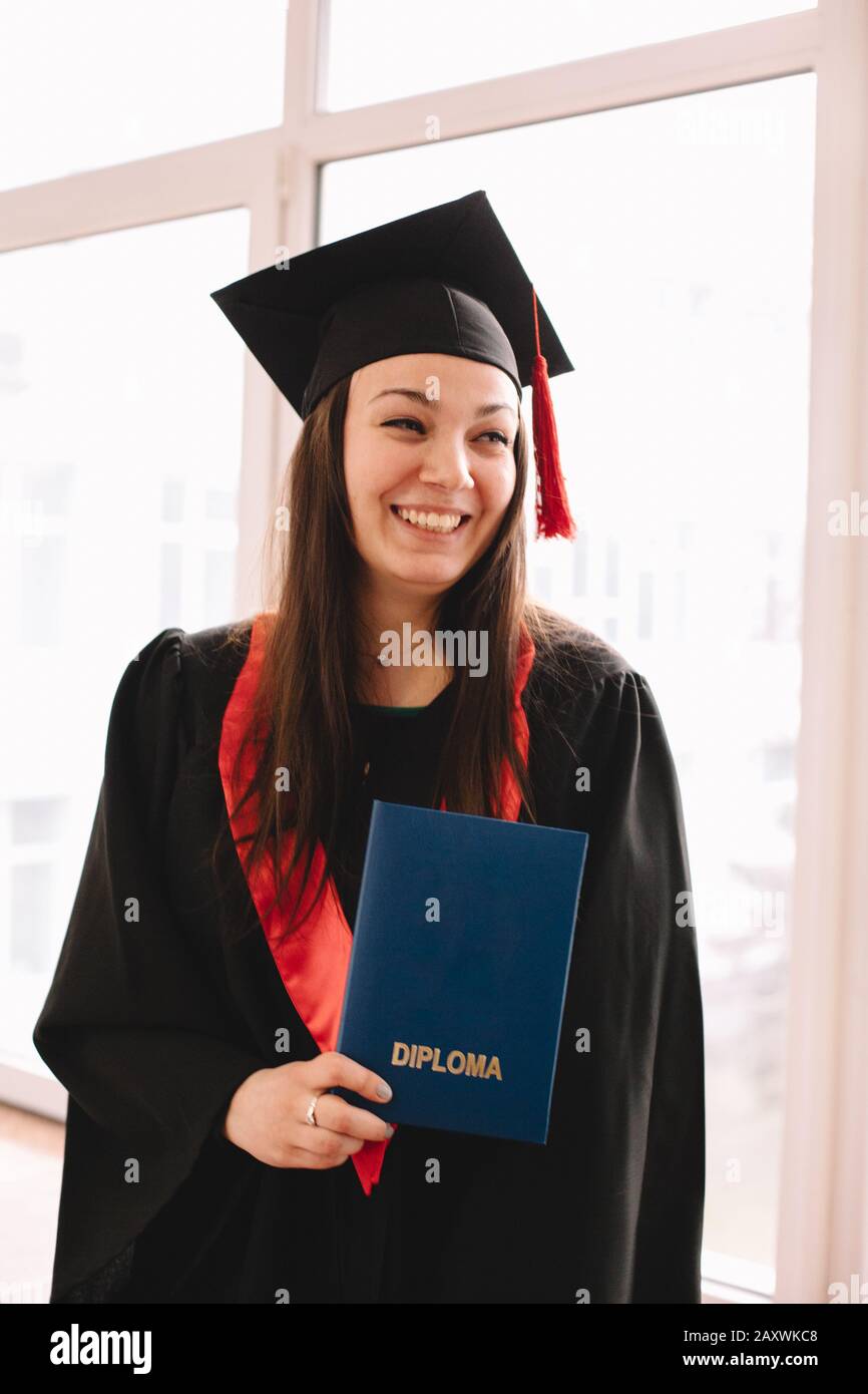 Young cheerful female graduate student wearing mortarboard holding diploma while standing in university at graduation ceremony Stock Photo