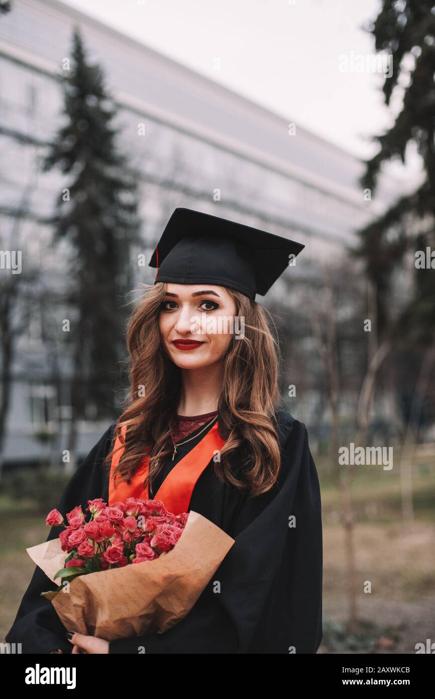 Portrait of young woman in graduation gown holding bouquet of flowers