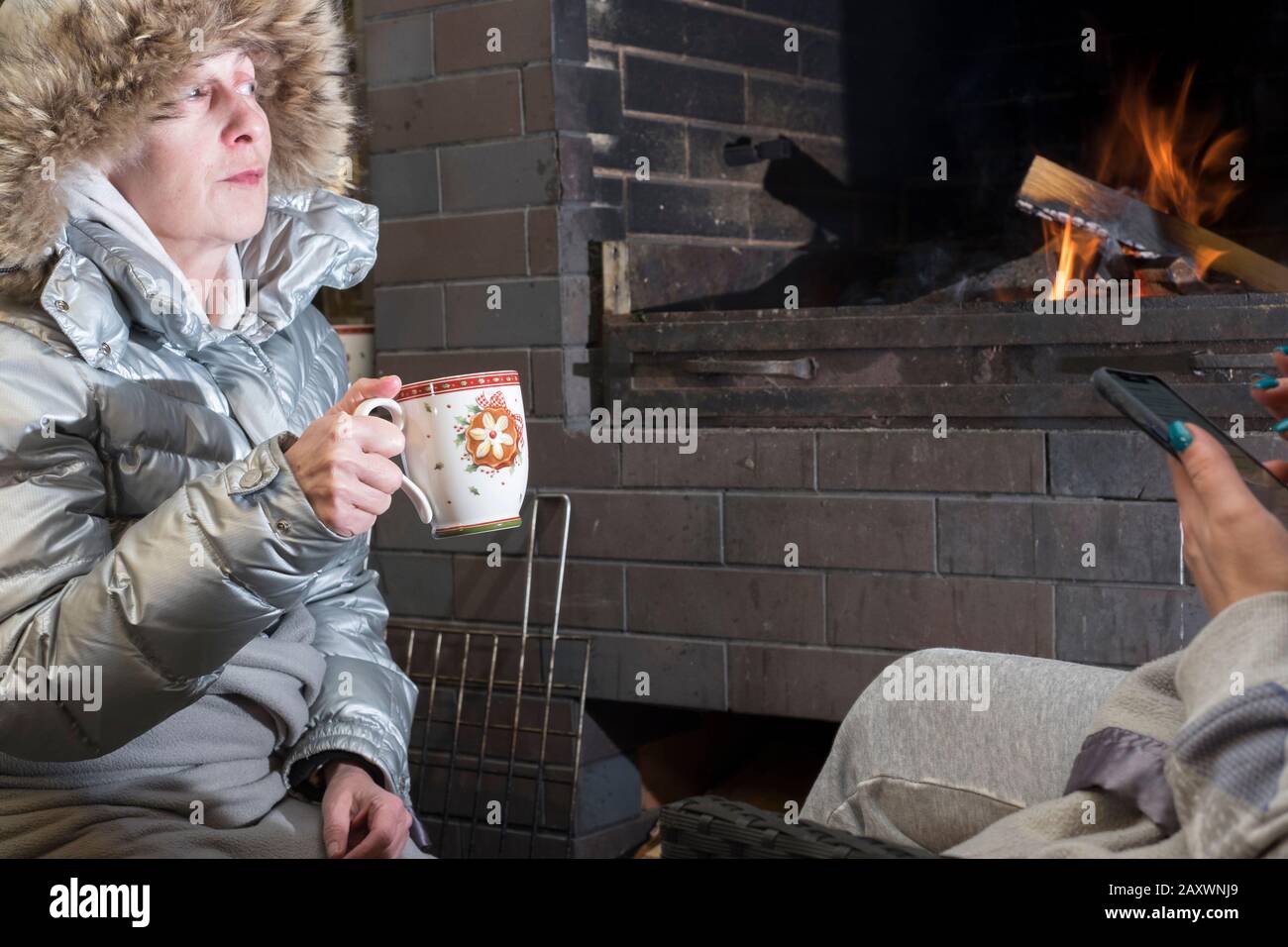 One woman drinking from a mug next to a fireplce, another texting on mobile, putdoot night scene Stock Photo