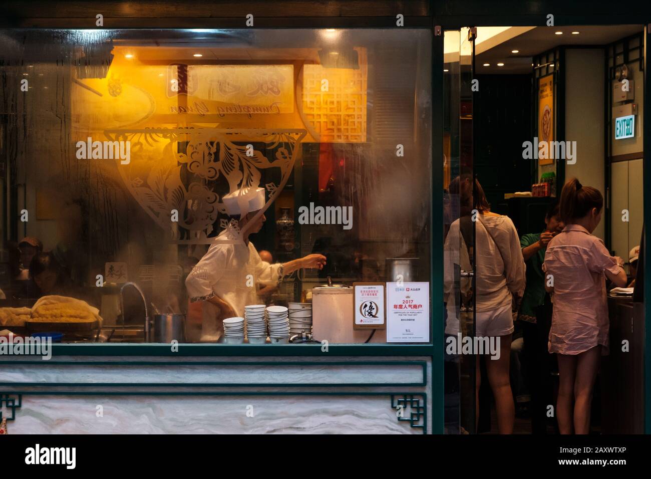 Hong Kong - November, 2019:  Window of Chinese restaurant showing chef cooking food and people eating Stock Photo