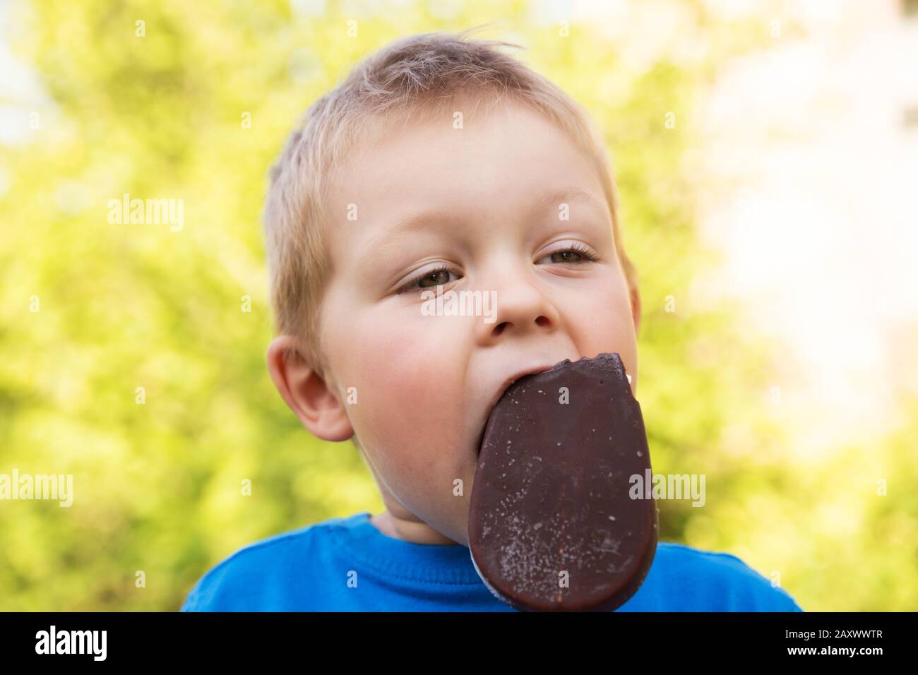 Attractive blond boy of 5 years in the summer outdoors on a hot sunny day eating popsicle in chocolate on a stick. Closeup portrait on blurry backgrou Stock Photo