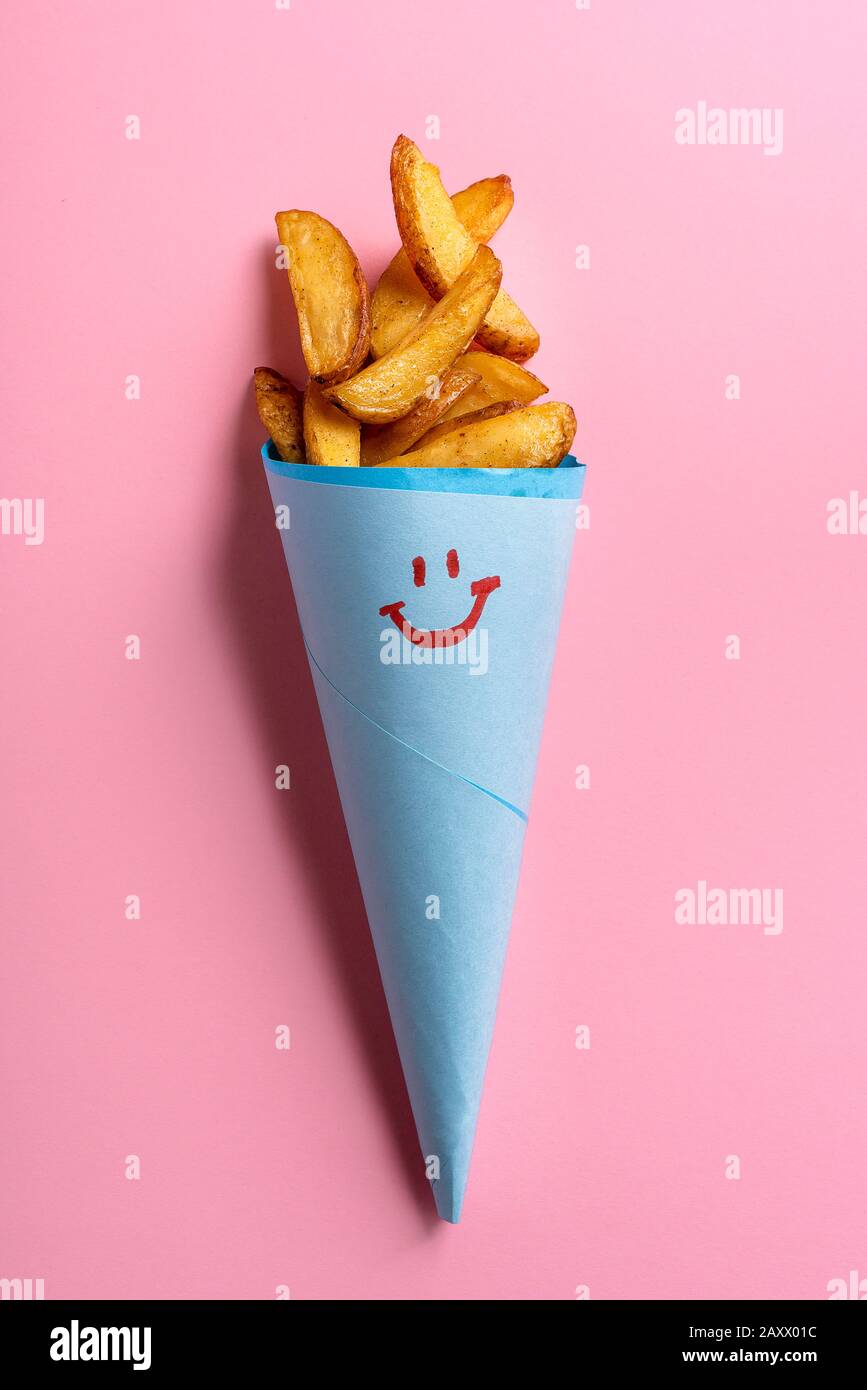 Baked potatoes wedges in a blue paper cone with a smiley face on a pink background. Tasty roast potatoes in a cone. Homemade rustic potatoes. Stock Photo