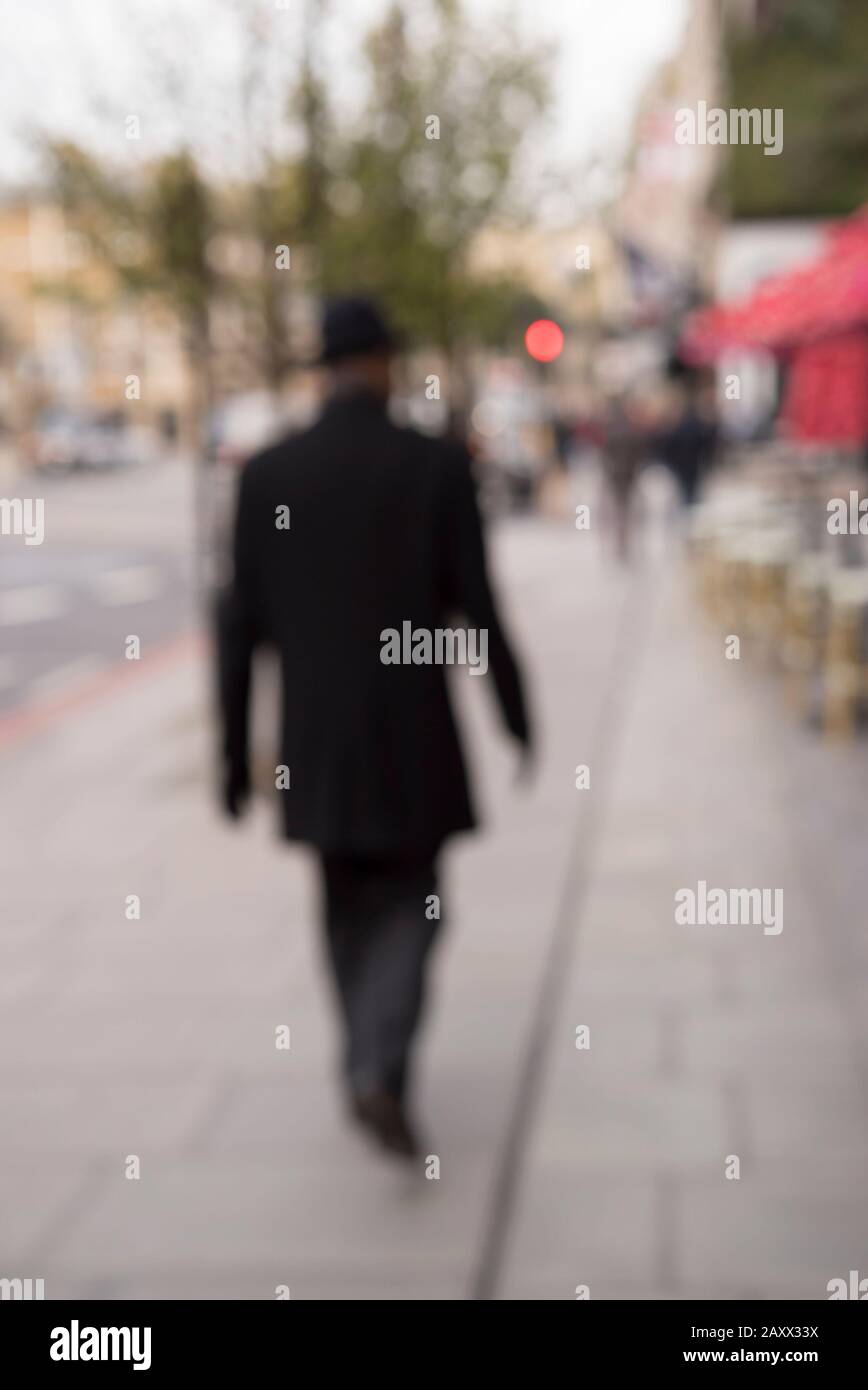 Rear view of mysterious blurred figure in black hat & coat, London street walking away on pavement. Strange mystery man, secret agent, undercover spy. Stock Photo