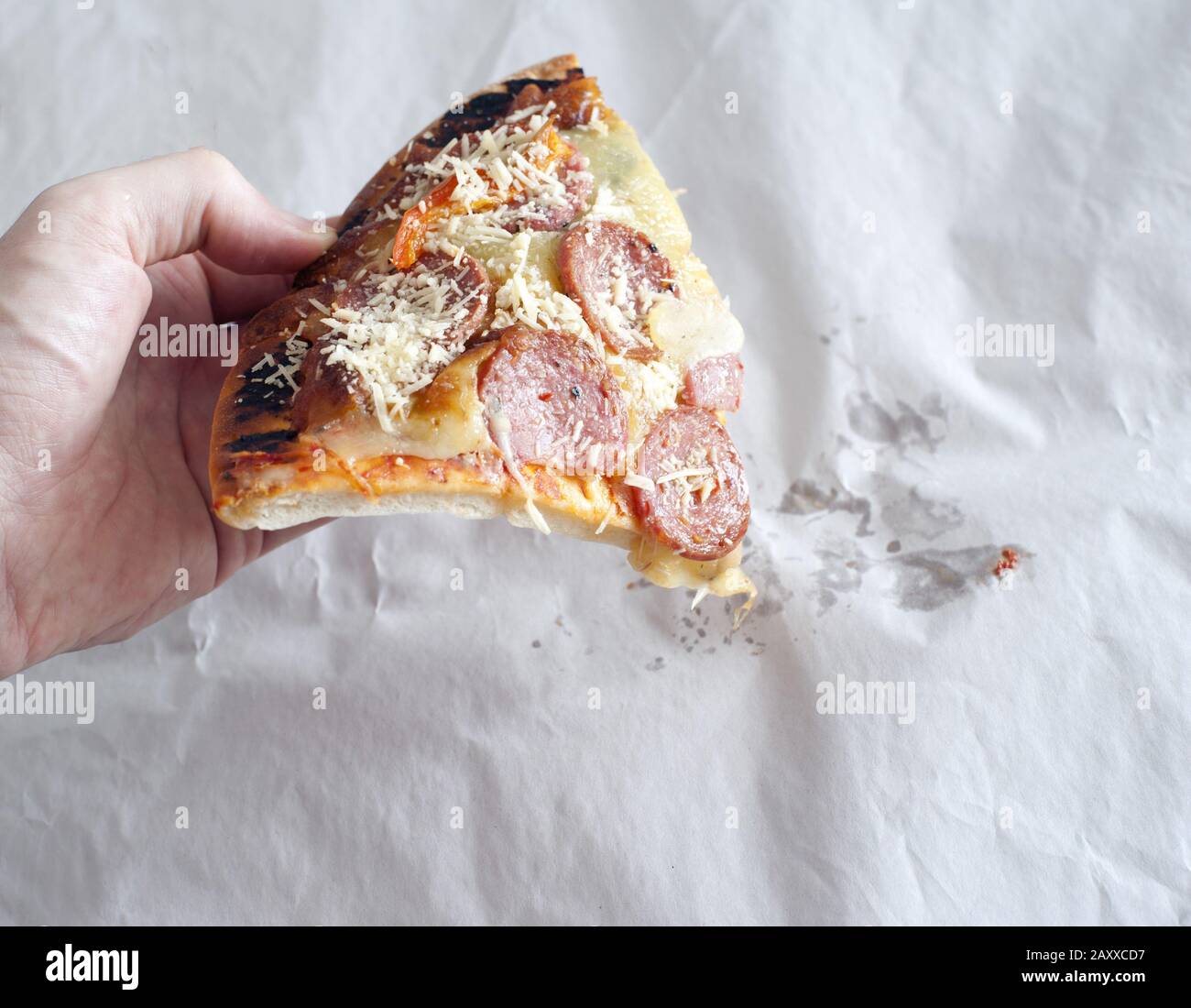 Hand holding greasy unappealing pepperoni pizza slice over white wrapping paper with grease