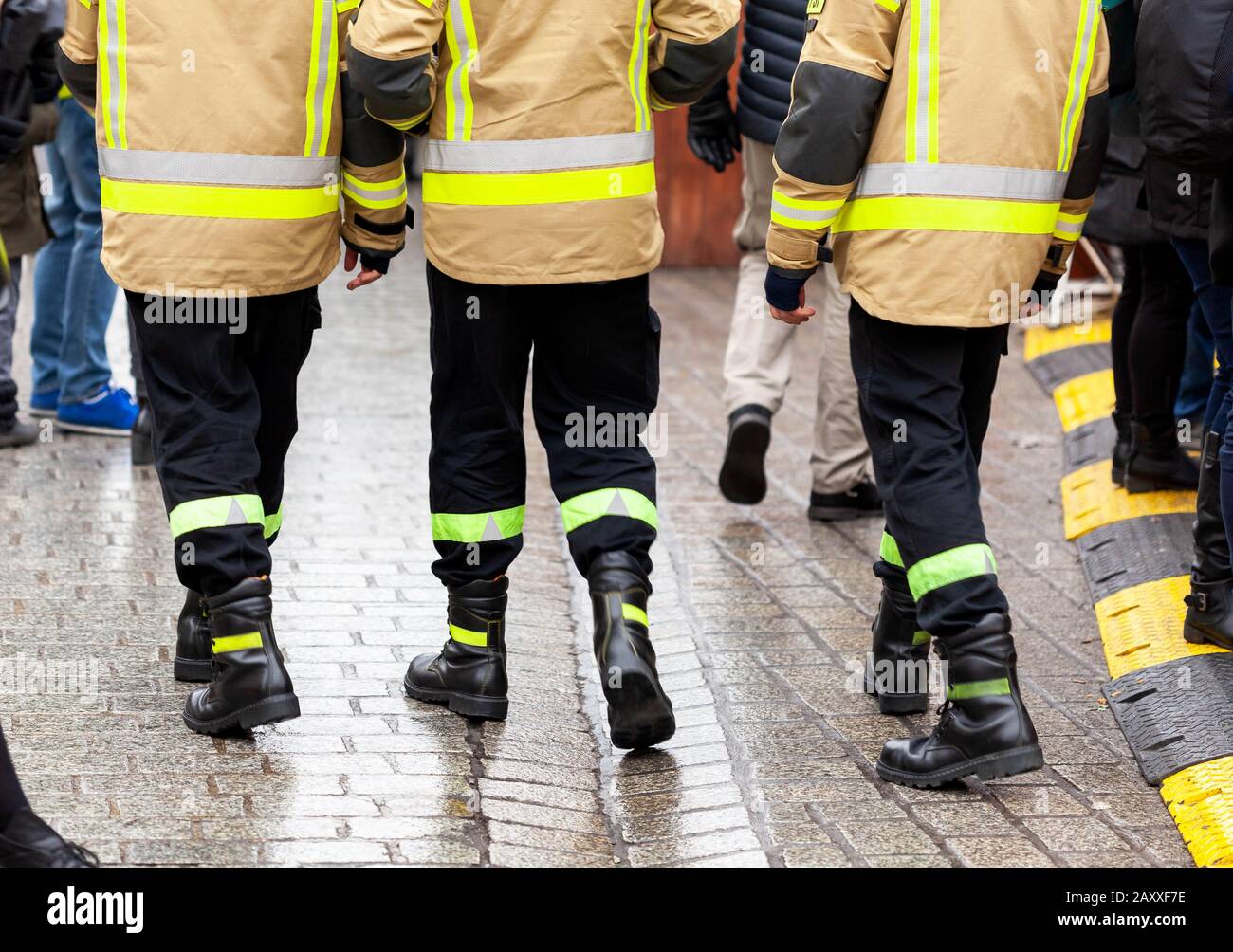 Three anonymous fireman in yellow reflective uniforms and black boots and trousers walking away from the camera on the crowdy street, back side, legs Stock Photo