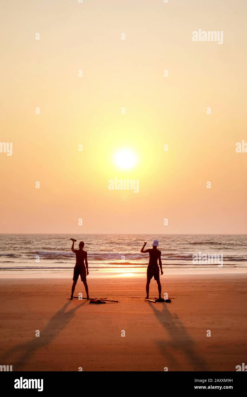 two unrecognizable men exercising at sunset by holding two small weights on a wide open empty sandy beach, they are standing with their arms outstretc Stock Photo