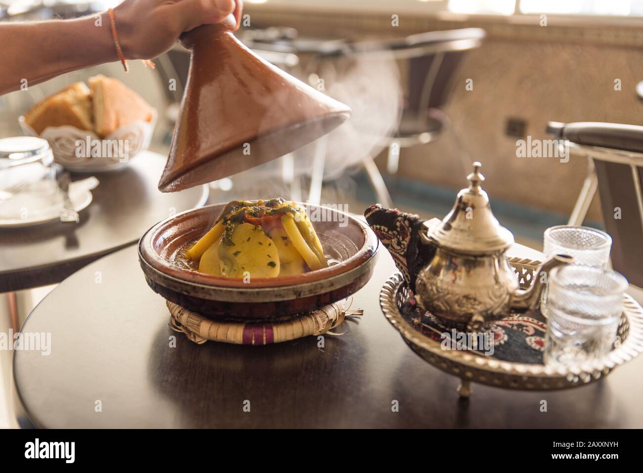Lifting the Lid off a Tagine in a Moroccan Restaurant With Traditional Teapot and Table Setting Stock Photo