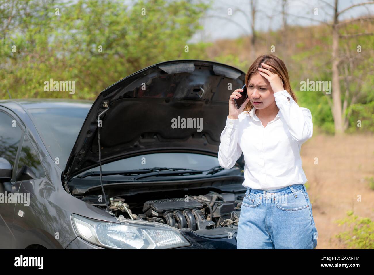 Asian young unhappy woman talking on a cell phone  in front of the open hood  broken down car On Country Road Phoning For Help. Broken Car On The Road Stock Photo