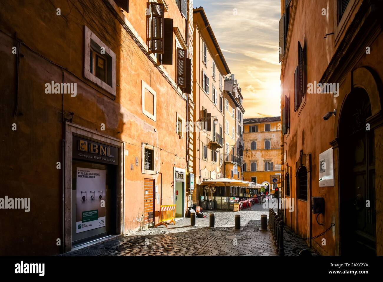 A narrow alley with high walls on either side leads to a small square with a sidewalk cafe as the sun sets in Rome, Italy. Stock Photo