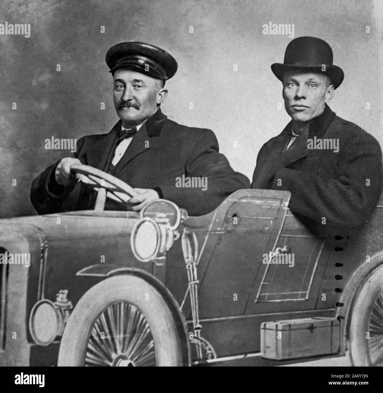 A dour  looking man poses with his 'chauffeur' behind an amusement park cut-out of a car, ca. 1905. Stock Photo